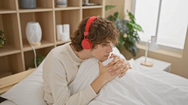 Kulaklıklı düşünceli genç bir adam, modern yaşam tarzını yansıtan, iyi ışıklandırılmış bir yatak odasında yalnızlıktan zevk alır..