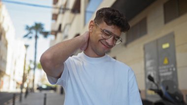 Gözlüklü ve beyaz gömlekli yakışıklı genç adam güneşli bir günde şehir parkında dikilirken rahatsız bir şekilde boynunu ovalıyor..