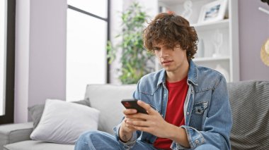 Çağdaş bir oturma odasında gri bir koltukta otururken telefonda mesaj atan, kot pantolonlu kıvırcık saçlı genç bir adam..
