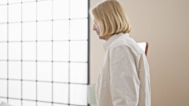 Beyaz tişörtlü, omuz hizasında sarı saçlı, düşünceli bir adam doğal ışık alan tarafsız bir zemine karşı içeride duruyor..