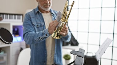 A senior man cleans a trumpet in a modern office with musical equipment. clipart