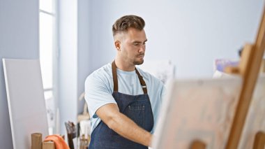 Sakallı, kot önlüklü genç bir İspanyol, parlak bir sanat stüdyosundaki tuvale odaklanıyor..