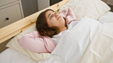 Düşünceli genç bir İspanyol kadın rahat bir yatak odasında uzanıyor, düşünceli ve rahat görünüyor..