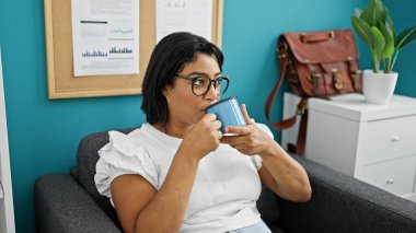 İspanyol olgun kadın modern bir ofiste kahve içiyor, gözlük takıyor ve dalgın görünüyor..