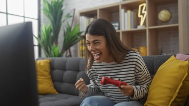 Excited hispanic woman playing video game in modern living room clipart