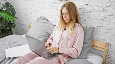 Pembe pijamalı genç sarışın bir kadın laptopuyla modern bir yatak odasında beyaz tuğlalı bir duvarda çalışıyor..