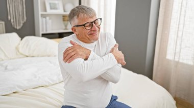 Gözlüklü, gülümseyen orta yaşlı bir adam rahat bir yatak odasında kendini kucaklıyor..