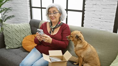 Gri saçlı olgun bir kadın, rahat bir oturma odasında nazik köpeğinin yanında akıllı telefonuyla etkileşime girer..
