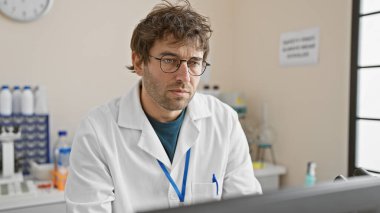 Gözlüklü ve sakallı laboratuvar önlüğü giyen odaklanmış genç bir adam hastane laboratuarında veri analiz ediyor..