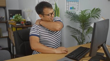 Genç yakışıklı Asyalı bir adam ofiste sırt ağrısı çekiyor. Arka planda bilgisayar, bitki ve ofis malzemeleriyle dolu bir masada otururken çizgili gömlek ve gözlük takıyor..