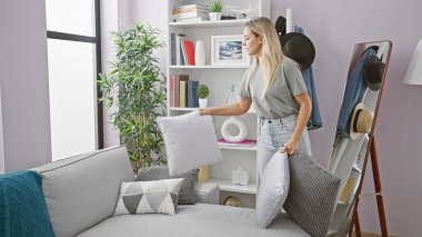 Genç sarışın bir kadın, günlük yaşantısını yansıtan şık ve modern bir oturma odasında yastık düzenliyor..