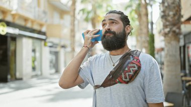 Yakışıklı sakallı İspanyol adam güneşli bir şehirde akıllı telefon kullanıyor.