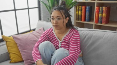 Düşünceli genç bir İspanyol kadın rahat bir oturma odasında, yastıklarla ve kitaplıkla çevrili bir koltukta oturuyor..