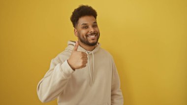 Gülümseyen Afrikalı Amerikalı adam sarı arka planda bej kapüşonlu başparmağını kaldırıyor.