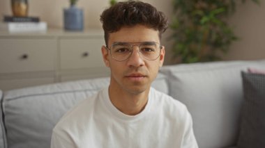 Genç, yakışıklı İspanyol adam gözlük takıyor ve arka planda modern dekoru olan rahat bir oturma odasında oturuyor..