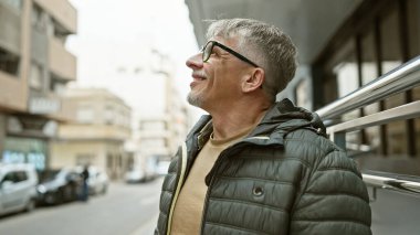 Gözlüklü, olgun, kır saçlı bir adam, bir şehir caddesinde düşünceli bir şekilde dikilirken kurnazca gülümsüyor, şehir hayatını anımsatıyor..