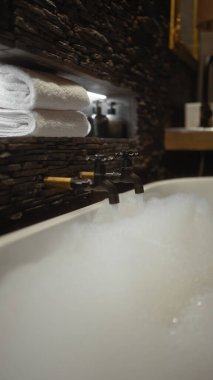 Köpüklü köpükle dolu lüks bir küvet, beyaz havlular ve dokulu taş bir duvara karşı lüks tuvaletler..