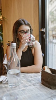 Gözlüklü esmer bir kadın modern bir kafede su içiyor ve sıradan bir yemek deneyimi sergiliyor..