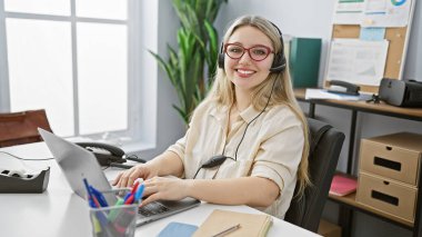 Modern ofis ortamında masasında çalışan, kulaklık takan, gülümseyen sarışın bir kadın.