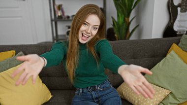 Yeşil kazaklı neşeli genç bir kadın gri bir koltukta oturuyor ve modern bir oturma odasında neşeyle elini uzatıyor..