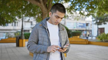 Şehir parkında arka planında yeşillik olan akıllı telefon kullanan genç bir İspanyol. Odaklanmış ve sıradan görünüyor..