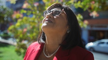 Kırmızı ceketli, kısa saçlı ve gözlüklü olgun bir İspanyol kadın şehir parkında güneşli bir günün tadını çıkarıyor..