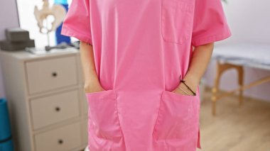 A female healthcare professional in pink scrubs stands confidently in a medical clinic's examination room. clipart