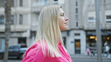 Pembe gömlekli genç sarışın bir kadın hareketli bir sokak köşesinde düşünceli bir şekilde duruyor, şehir güzelliğini ve günlük tarzını somutlaştırıyor..