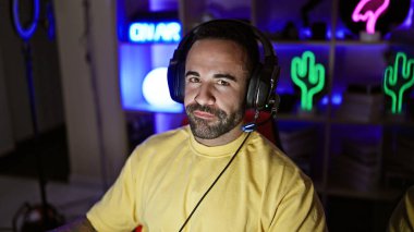 Gece vakti neon ışıkları olan bir oyun odasında sakallı, kulaklık takan genç İspanyol bir adam..