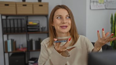 Genç, güzel, beyaz bir kadın ofiste otururken gülümsüyor ve telefonda konuşuyor..