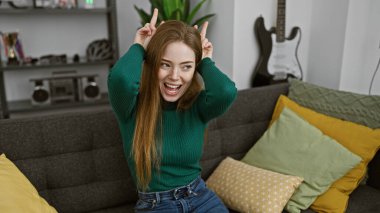 Modern dekorlu rahat bir oturma odasında komik surat ifadeleri takınan neşeli genç bir kadın.