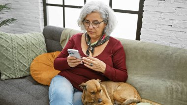 Gözlüklü olgun bir kadın, oturma odasının iç kısmında kanepede köpeğiyle akıllı telefon kullanıyor..