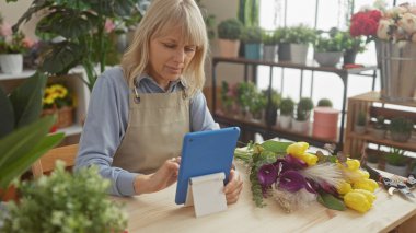 Çiçekçide tablet kullanan sarışın kadın. Etrafı renkli buketler ve bitkilerle çevrili..