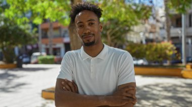 Güneşli bir günde şehir parkında, sakallı, yakışıklı, Afro-Amerikalı bir adam, güvenli bir şekilde şehir ortamında kollarını kavuşturuyor..