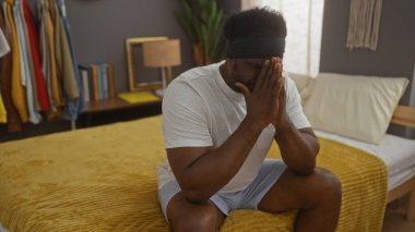 Genç Afro-Amerikalı bir adam yatak odasında, elleri sımsıkı bağlanmış bir şekilde oturuyor, rahat, modern bir ev içinde tefekkür ediyor..