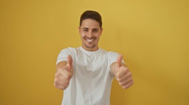 Sakallı, gülümseyen genç İspanyol adam sarı arka plana başparmak sallıyor..