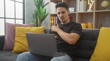Sakallı genç bir adam rahat ve modern bir oturma odasında dizüstü bilgisayarla çalışıyor..