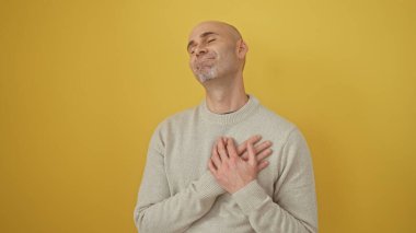 Sakallı, sakallı, sarı arka planda duran ve kalbini tutan genç, kel bir adam..
