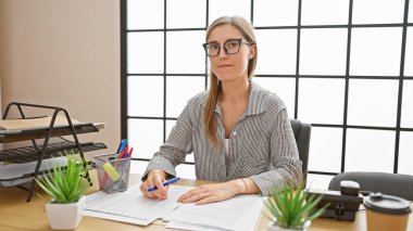 Tipik iş yeri eşyalarıyla çevrili modern ofis ortamında özenle çalışan odaklanmış beyaz kadın..