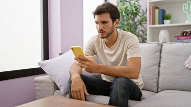 Evdeki kanepede otururken akıllı telefon kullanan yakışıklı İspanyol bir adam. Sıradan bir yaşam tarzı ve bağlantı hissi yayıyor..