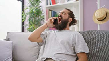 Sakallı ve sıradan saç stili esneme hareketleri olan genç bir İspanyol adam evde bir kanepede akıllı bir telefonla konuşuyor.
