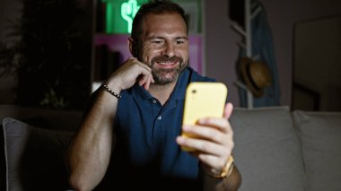 Yakışıklı, beyaz saçlı olgun bir adam gecenin karanlığında karanlık bir oturma odasında akıllı telefonuna gülümsüyor.