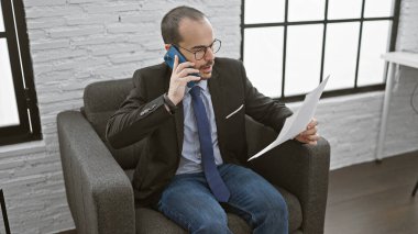 İş elbisesi içinde sakallı, telefonda konuşan ve ofis ortamında evrak okuyan İspanyol kel bir adam..