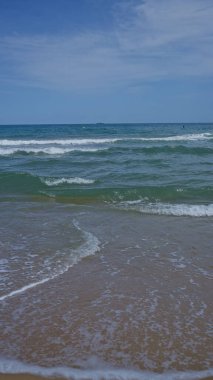 Serin mavi okyanus dalgaları parçalı bulutlu bir gökyüzünün altında kumlu bir plaja doğru yavaşça süzülerek kıyı manzarasının sükunetini yakalıyor..