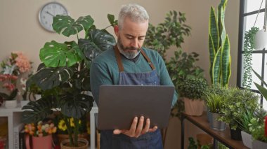 Mature man with beard using laptop in a plant-filled flower shop, portraying a small business owner or florist. clipart