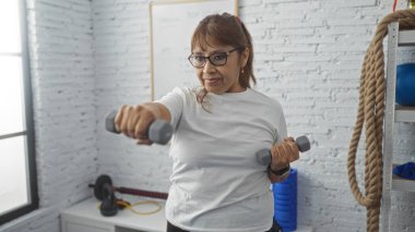 Orta yaşlı İspanyol bir kadın spor salonunda halterlerle egzersiz yapıyor. Parlak ve düzenli bir spor salonu var..
