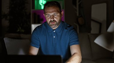 Beyaz saçlı, olgun İspanyol bir adam gecenin geç saatlerinde karanlık bir oturma odasında dizüstü bilgisayarla çalışıyor..