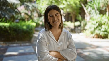 Genç ve güzel İspanyol kadın parkta kollarını kavuşturup gülümsüyor.