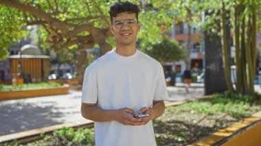 Güneşli bir günde, şehir parkında gülümseyen ve dışarıda akıllı bir telefon tutan İspanyol genç adam..
