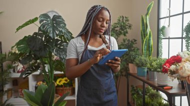 Saçları örgülü Afrikalı Amerikalı bir kadın yeşil bir çiçekçide tablet kullanıyor.